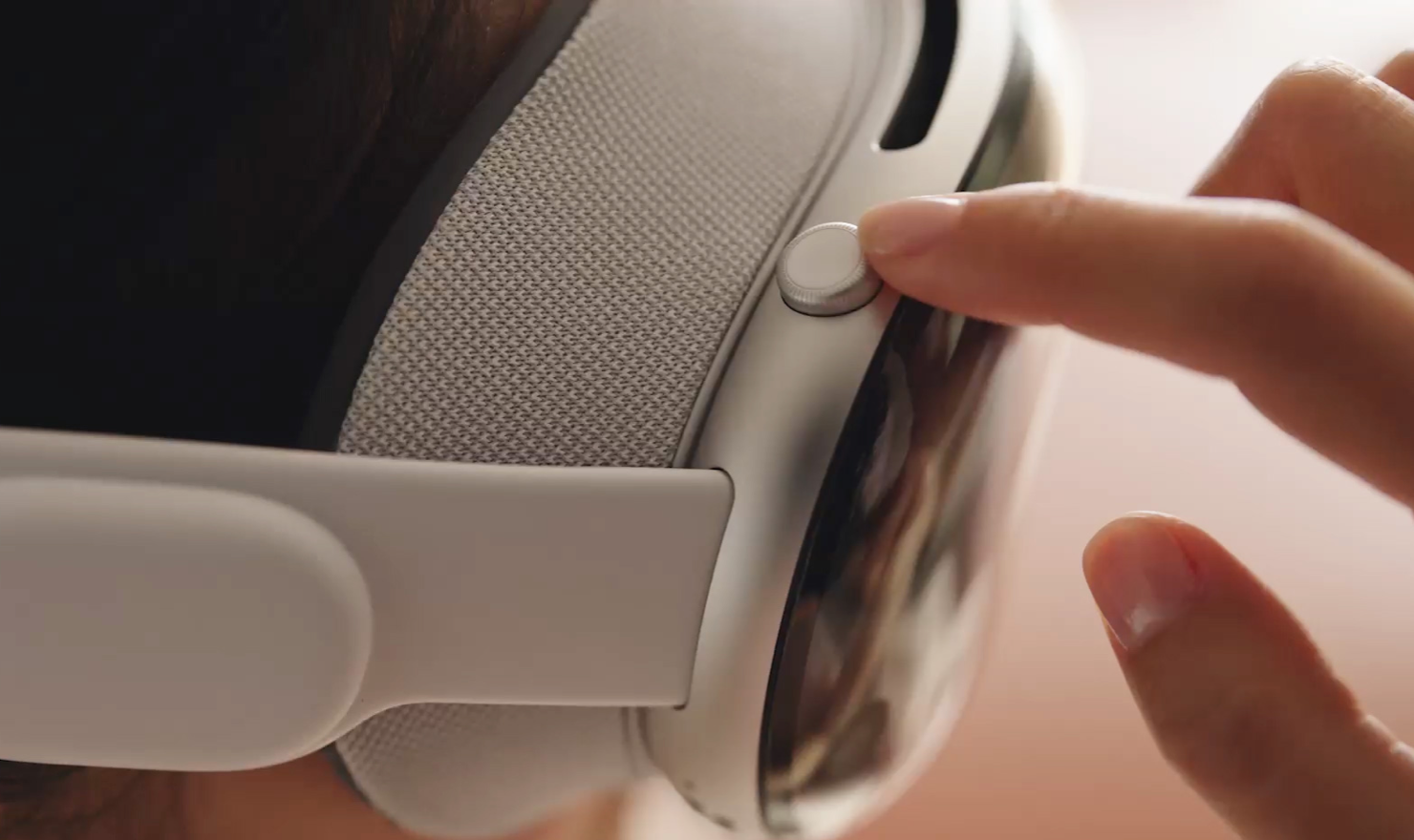 A close-up photograph of a person's head wearing Apple Vision Pro, with their index finger pointing at the Digital Crown.
